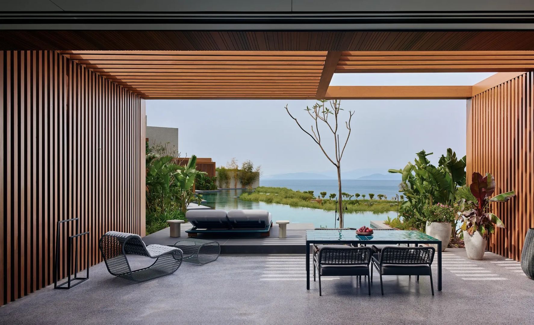 Überdachte Terrasse mit Sitzmöbeln, Esstisch und Blick auf einen Pool und das Meer im Hintergrund.