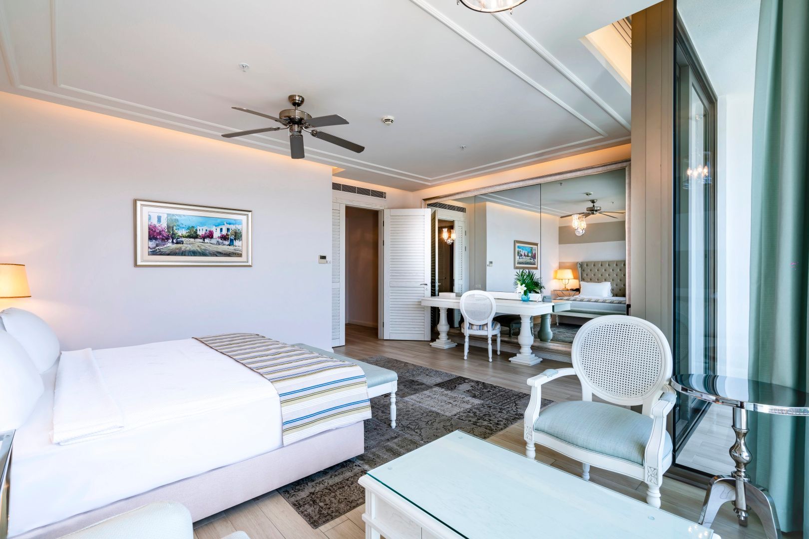 Helles Hotelzimmer mit großem Doppelbett, weißem Sitzbereich, Wandspiegel und Ventilator.