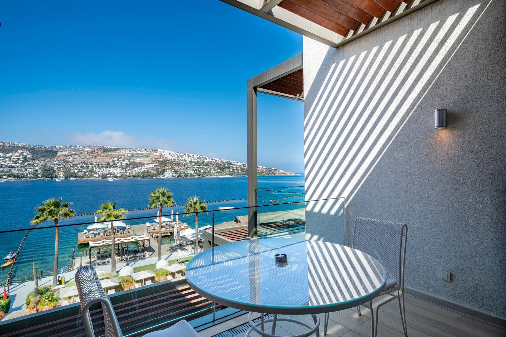 Blick von einem Balkon mit Glastisch und Stühlen auf das blaue Meer und die hügelige Küste.
