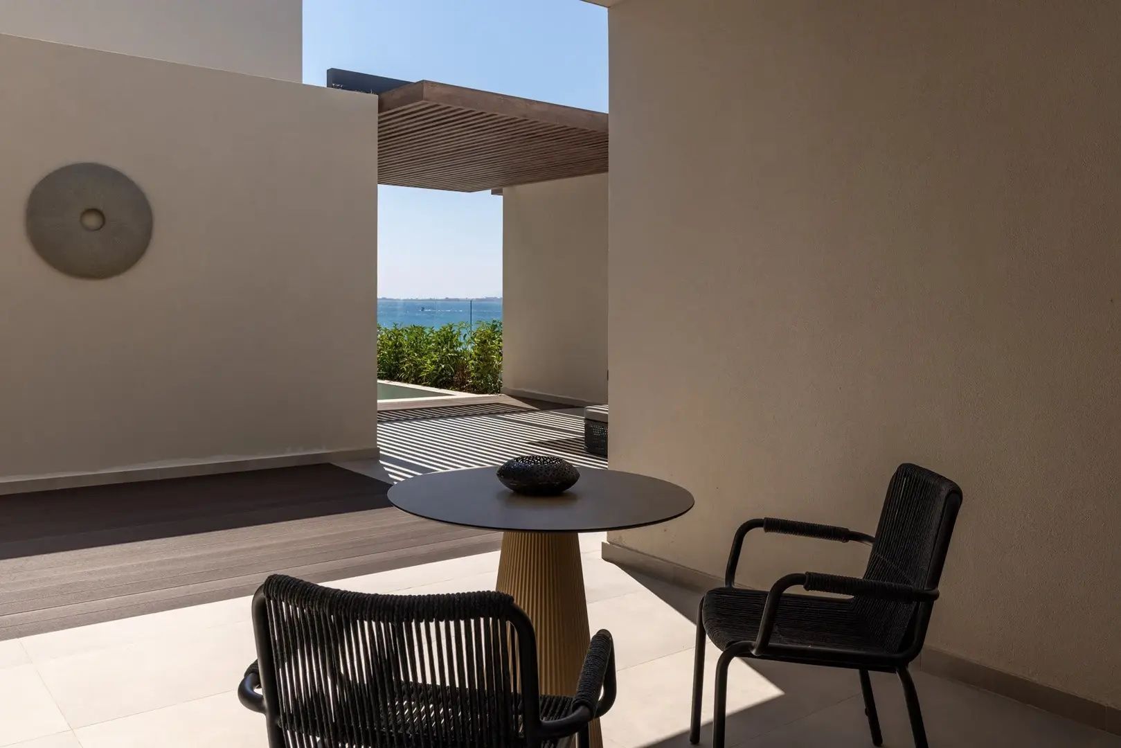 Überdachter Sitzbereich auf einer Terrasse mit rundem Tisch, zwei Stühlen und Blick zum Meer.