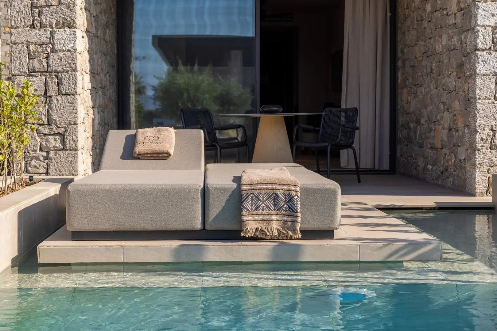 Gepolsterte Sonnenliegen auf der Terrasse direkt am Pool mit Sitzgruppe dahinter.
