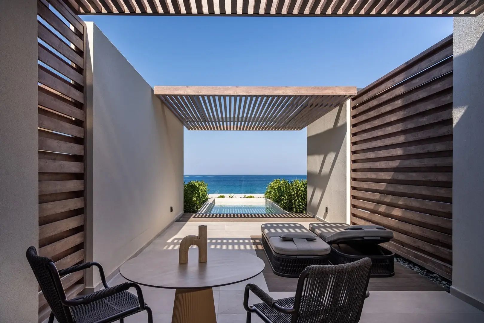 Überdachte Terrasse mit Sonnenliegen, Esstisch und Blick auf einen Pool und das Meer.