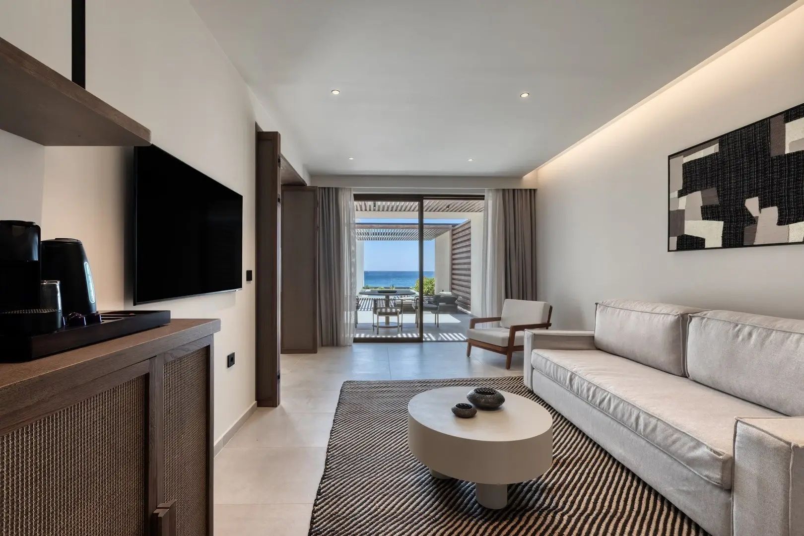 Hell eingerichteter Wohnbereich mit Sofa, Couchtisch, Fernseher und Terrassentür mit Meerblick.