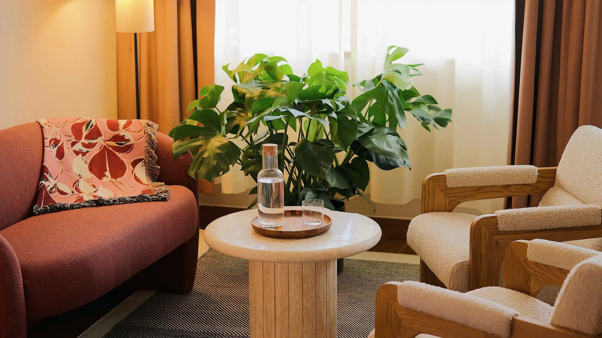 Runder Beistelltisch mit Wasserkaraffe vor einer großen Monstera und hellem Vorhang.