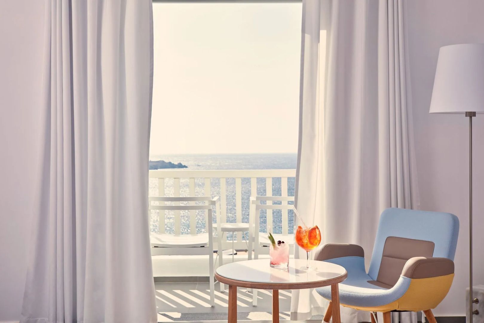 Blick durch die Balkontür auf zwei Getränke auf einem Tisch und das blaue Meer im Hintergrund.
