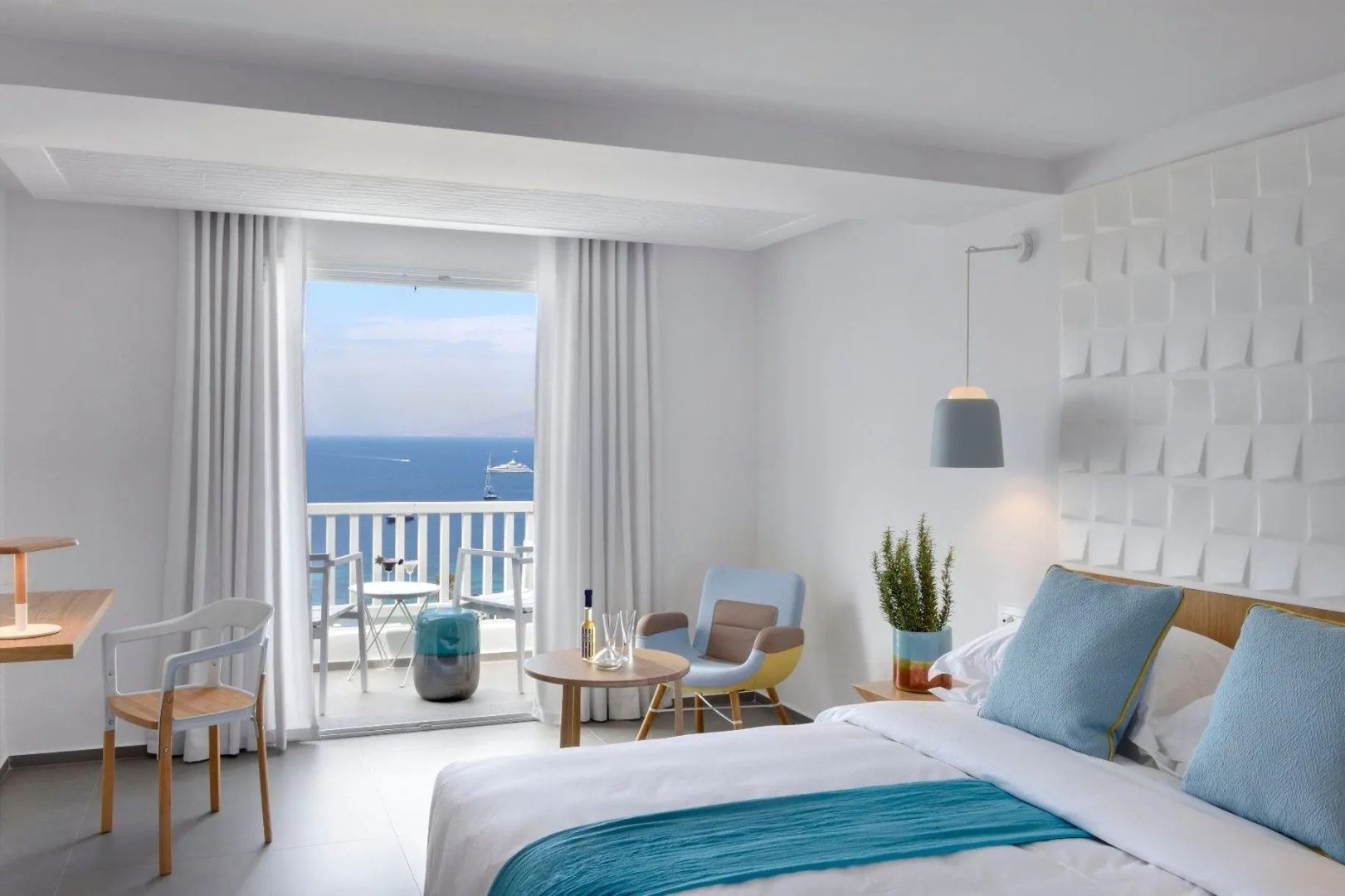 Hell eingerichtetes Hotelzimmer mit Doppelbett, Sessel und Blick auf das Meer durch die Balkontür.