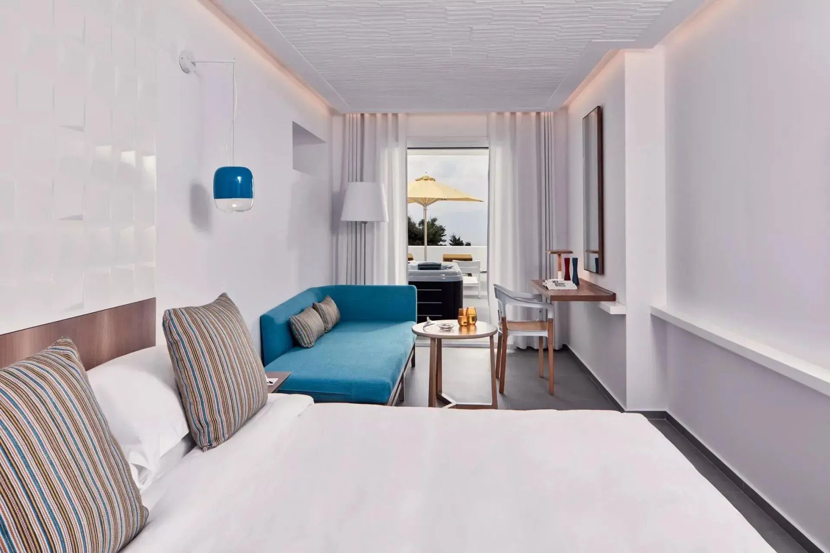 Hell gestaltetes Hotelzimmer mit blauem Sofa, Schreibplatz und Zugang zur privaten Terrasse.