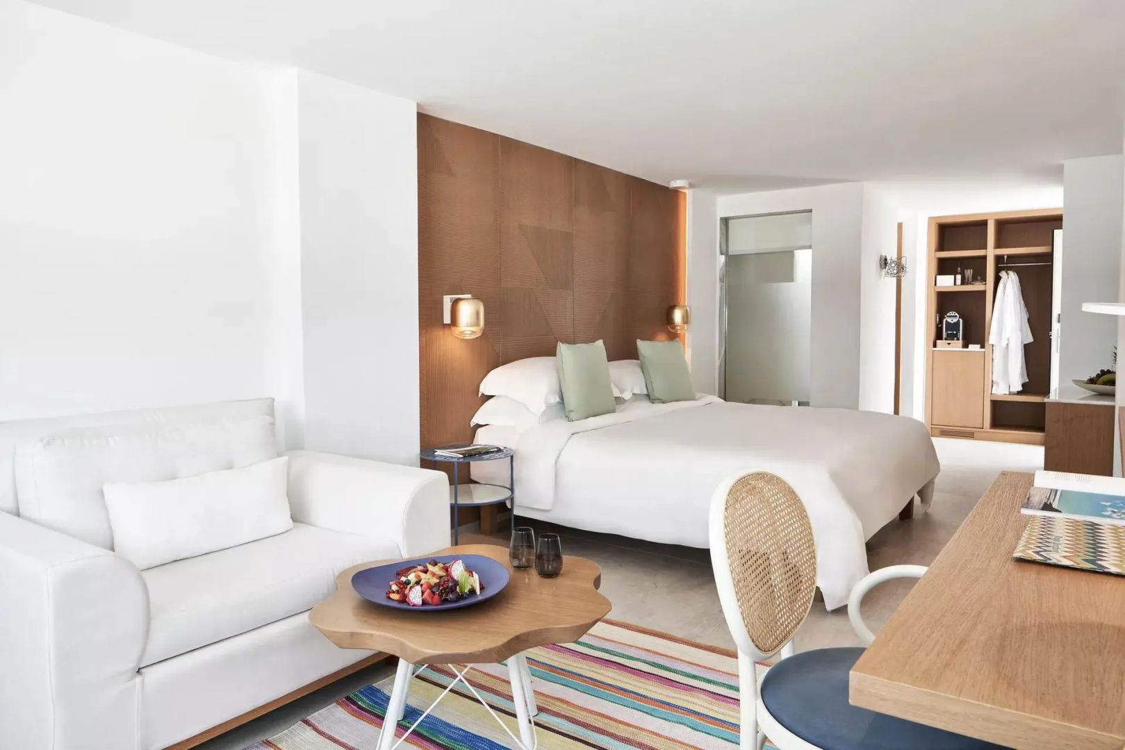 Doppelbett mit brauner Holzrückwand, weißem Sofa und buntem Teppich im Hotelzimmer.