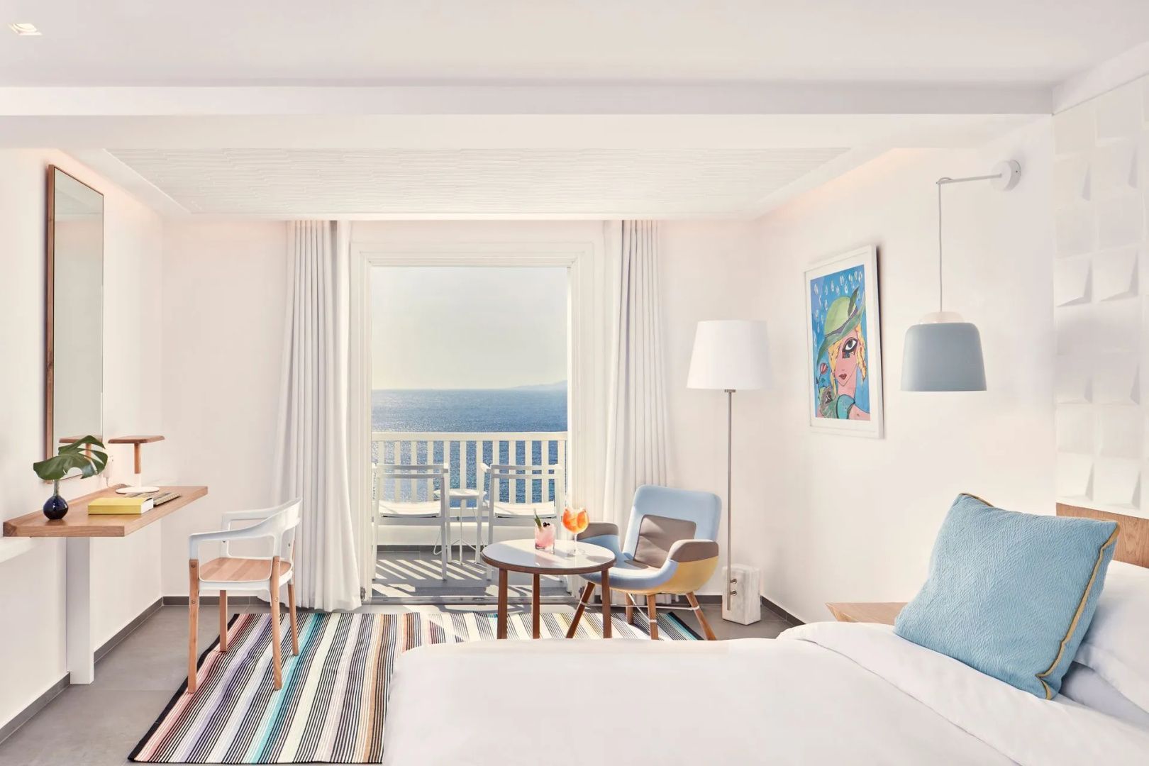 Weißes Hotelzimmer mit gestreiftem Teppich, Sitzgelegenheiten und Zugang zum Balkon mit Meerblick.