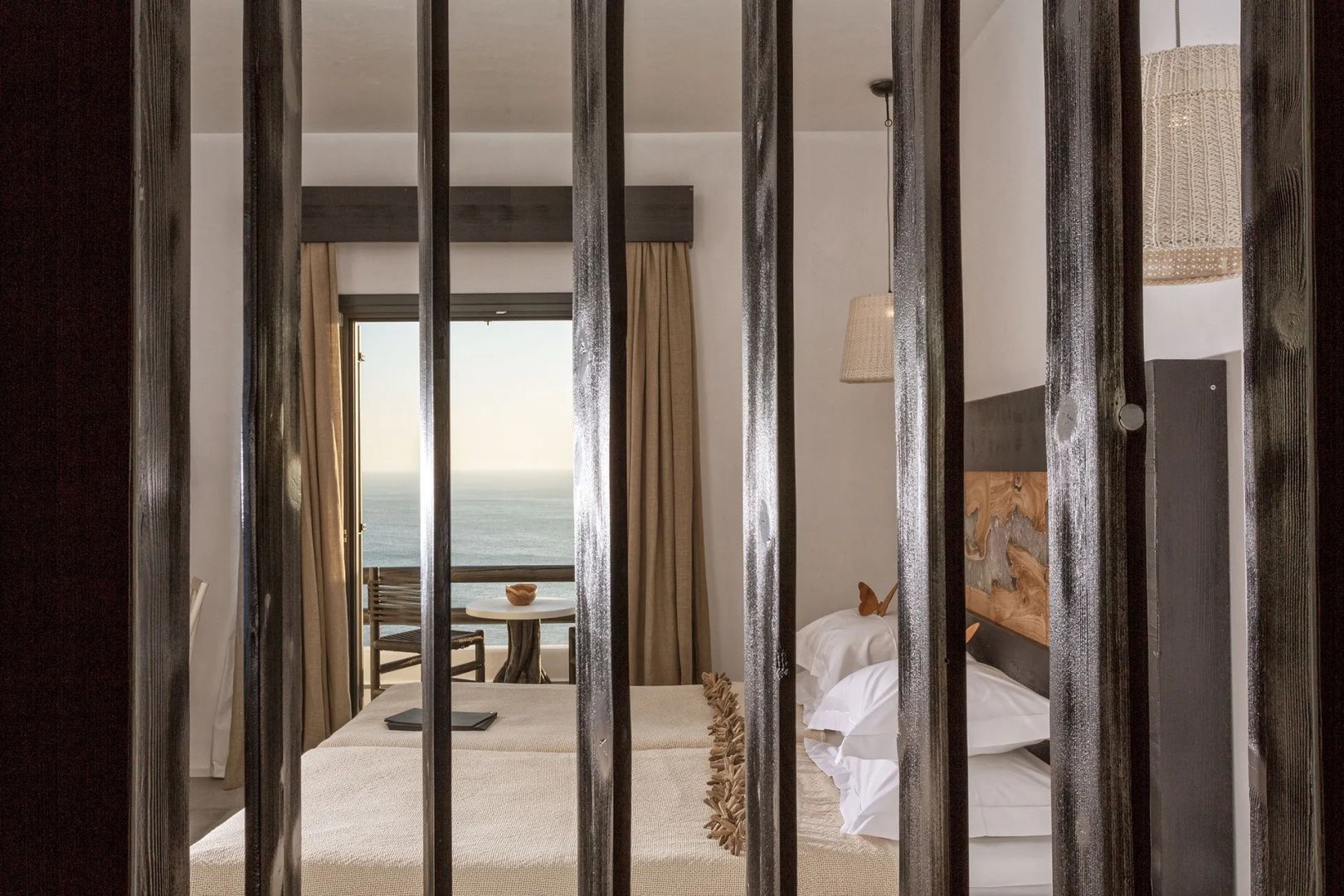 Blick durch Holzstreben auf ein Doppelbett und ein Fenster mit Blick auf das Meer.