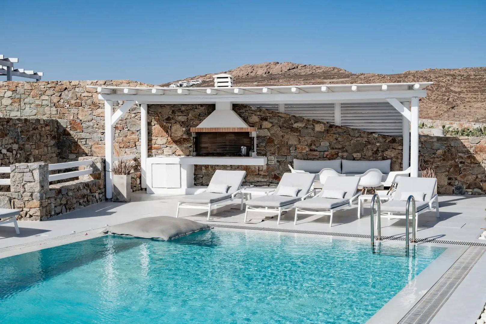 Türkisfarbener Pool mit weißen Sonnenliegen und einem gemauerten Grill unter einem Vordach.