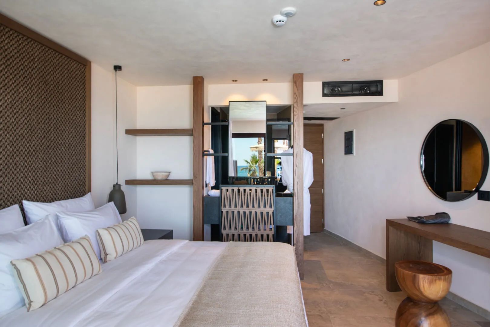 Helles Hotelzimmer mit Doppelbett, hölzerner Raumtrennung und offen gestaltetem Waschtischbereich.