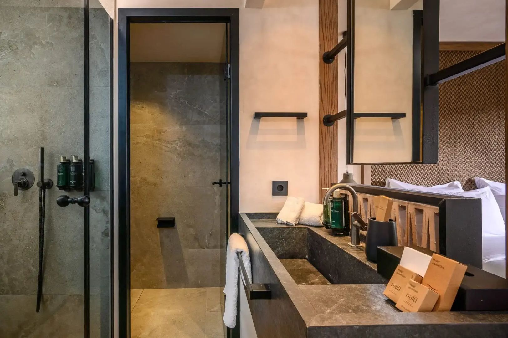 Badezimmer mit dunklem Waschtisch aus Stein, Spiegel, Pflegeprodukten und Dusche.