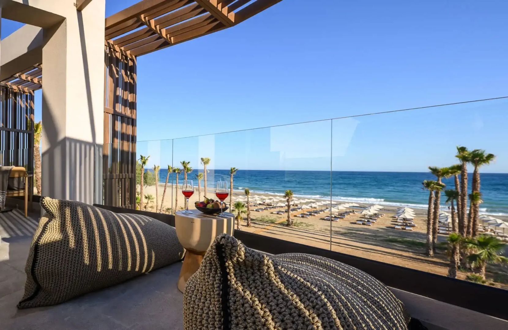Blick von einem Balkon mit Sitzkissen auf den Sandstrand, Palmen und das blaue Meer unter klarem Himmel.