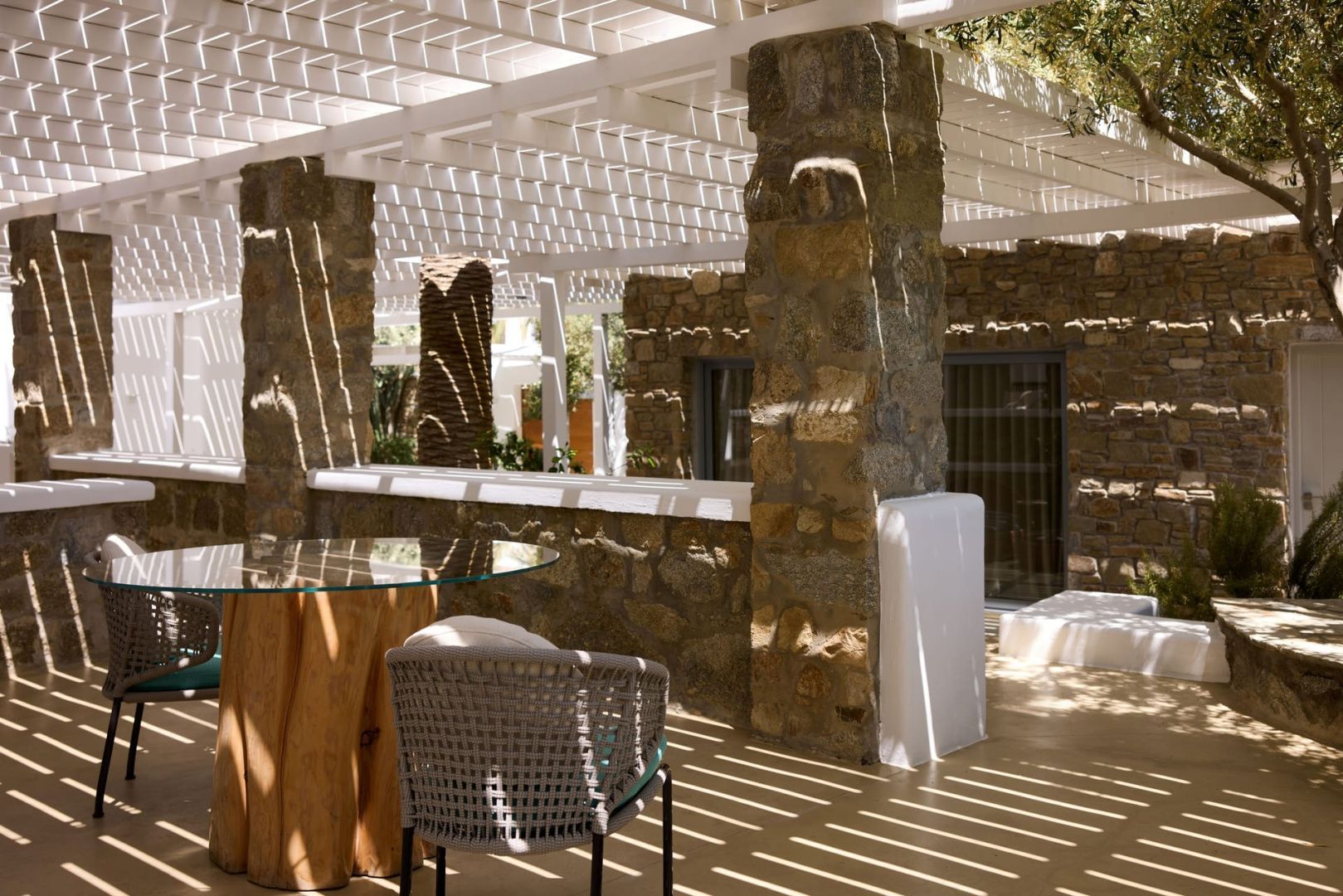 Außensitzplatz mit Holztisch und Flechtstuhl unter einer schattenspendenden Pergola.