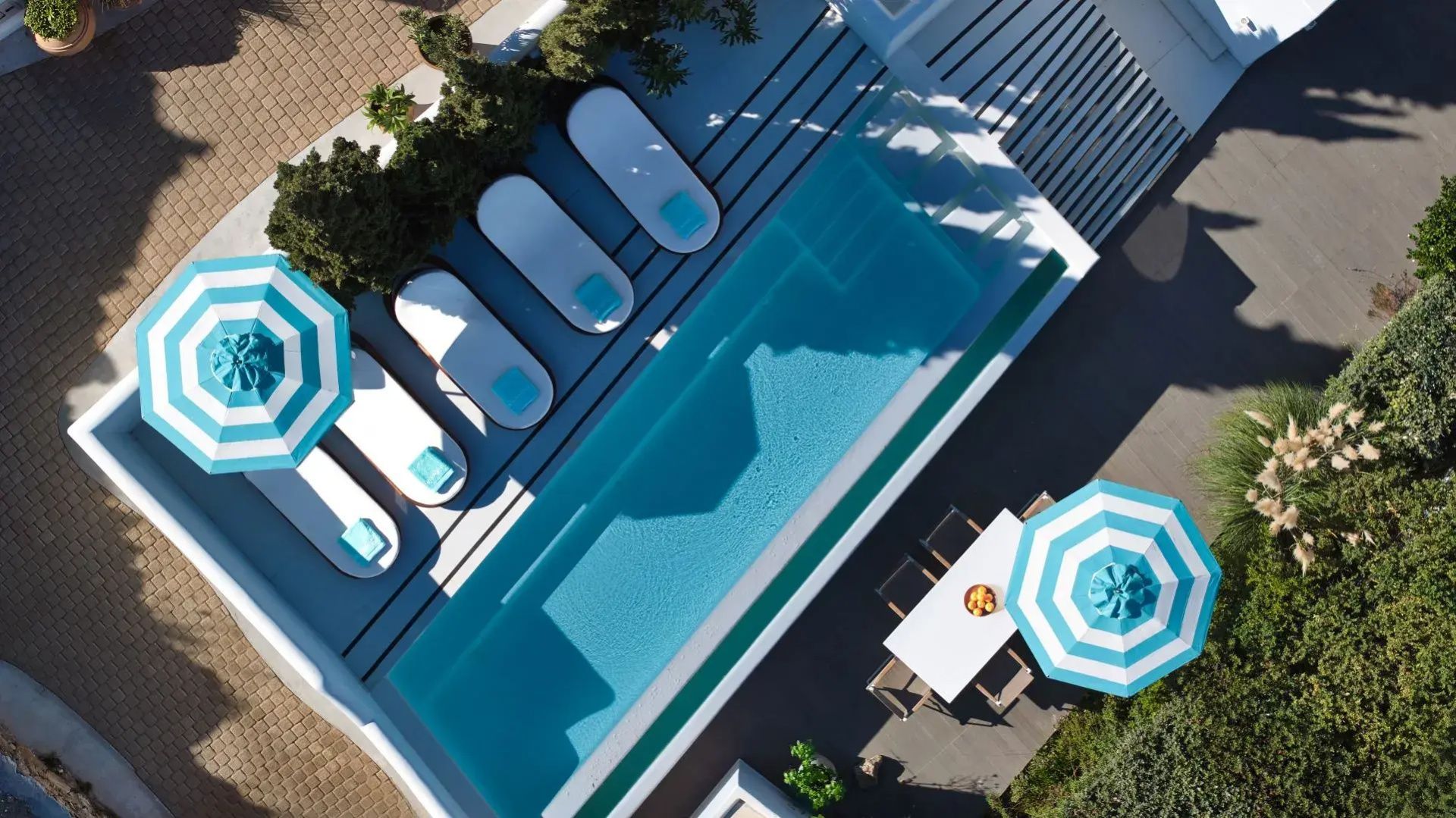 Blick von oben auf einen schmalen Pool mit weißen Sonnenliegen und Sonnenschirmen.