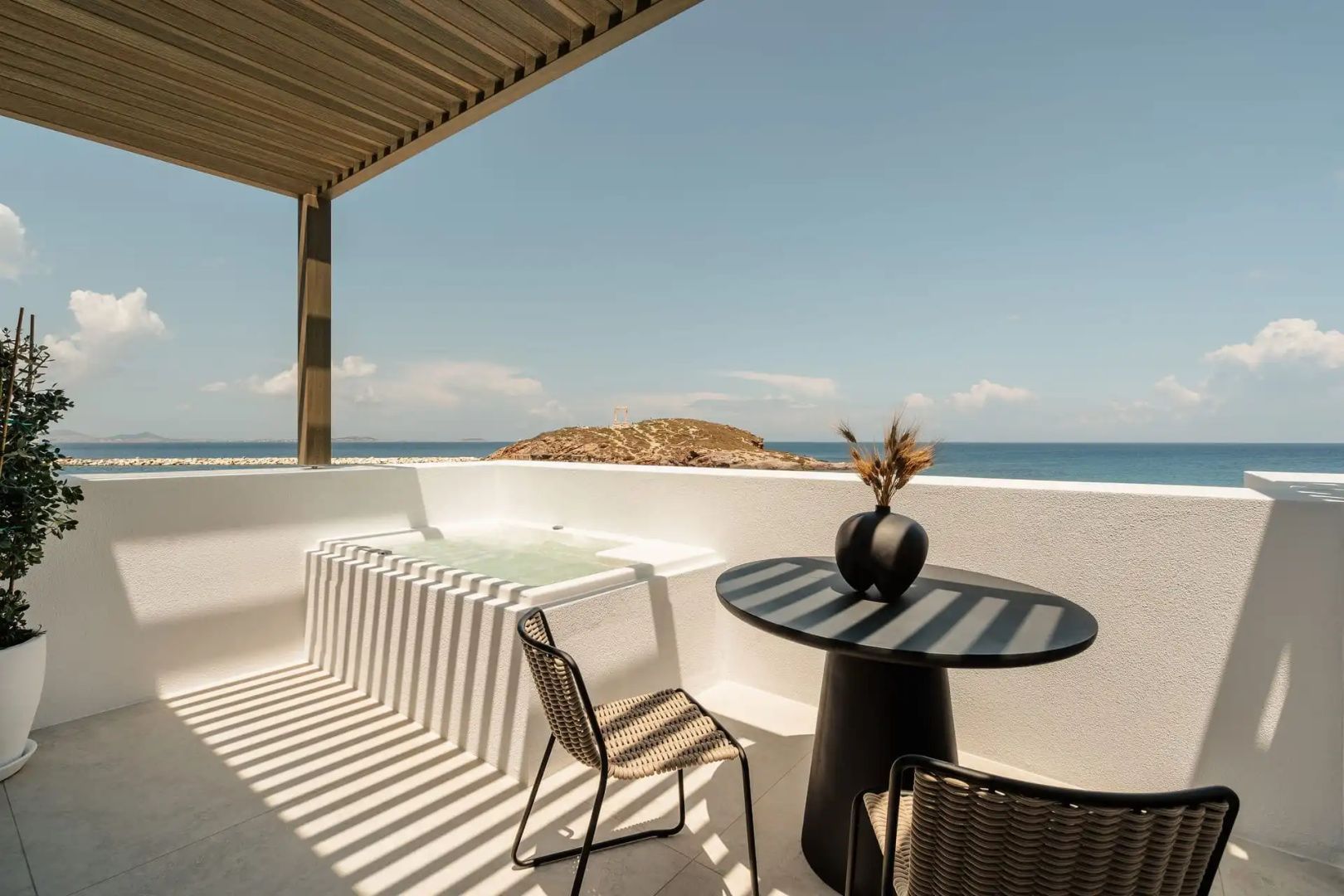 Sonnige Terrasse mit Whirlpool, Sitzgruppe und Weitblick auf eine Insel im Meer.