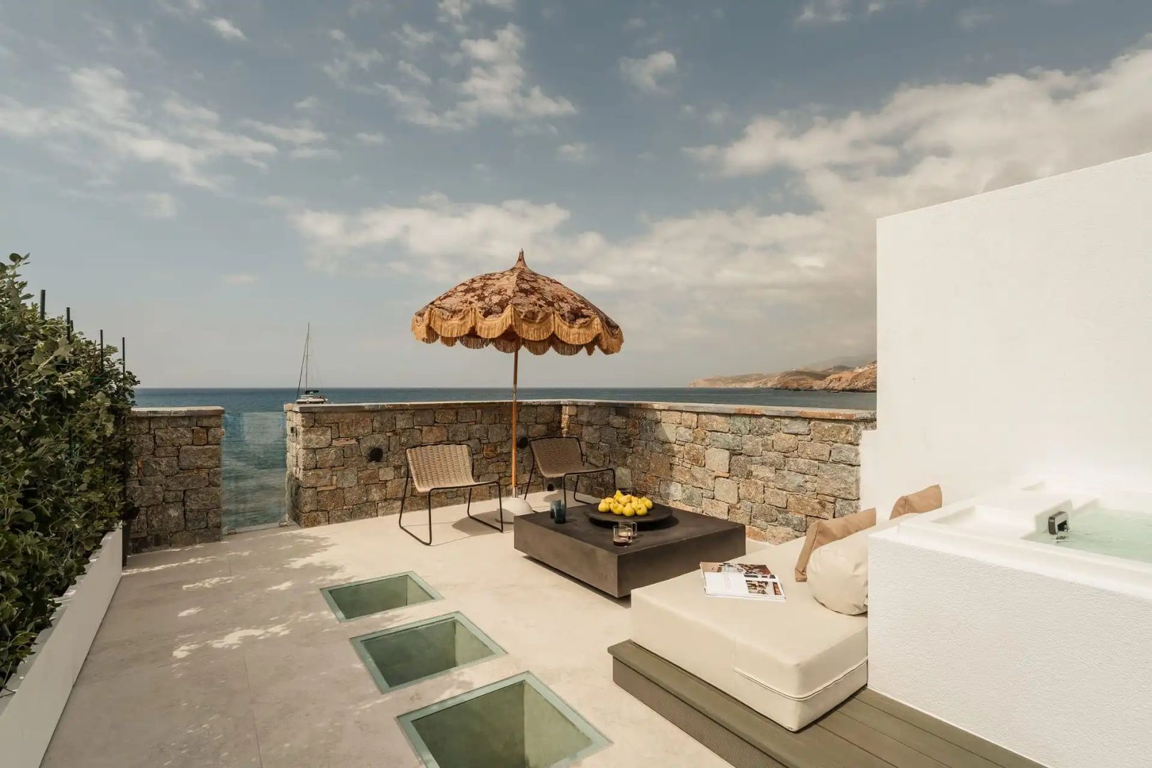 Terrasse mit braunem Sonnenschirm, Sitzgelegenheiten und Blick auf das blaue Meer.