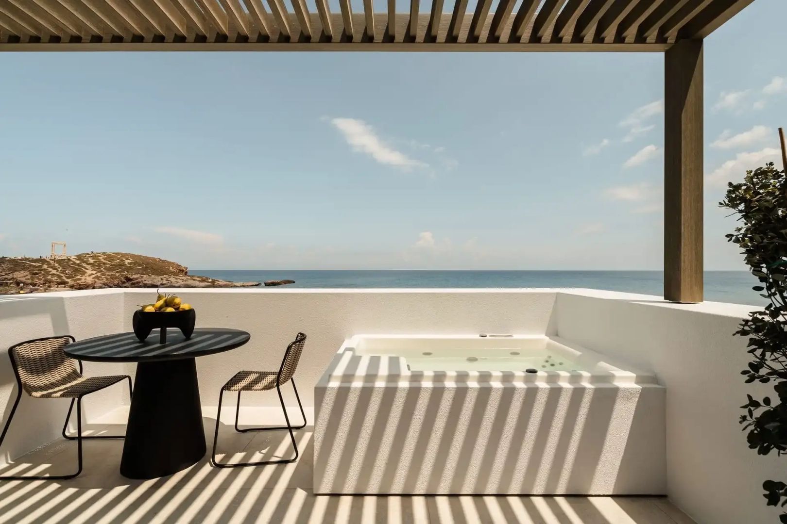 Terrassenbereich mit Jacuzzi, Esstisch und freiem Blick auf das blaue Meer unter sonnigem Himmel.