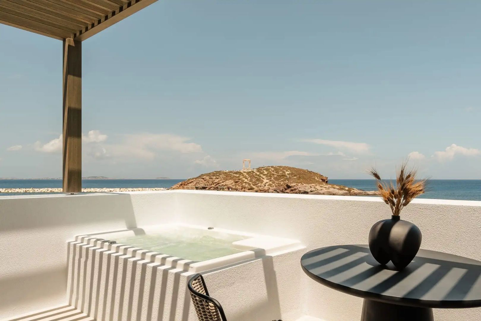 Blick von einer weissen Terrasse mit Whirlpool auf ein historisches Tor auf einer Felseninsel im Meer.