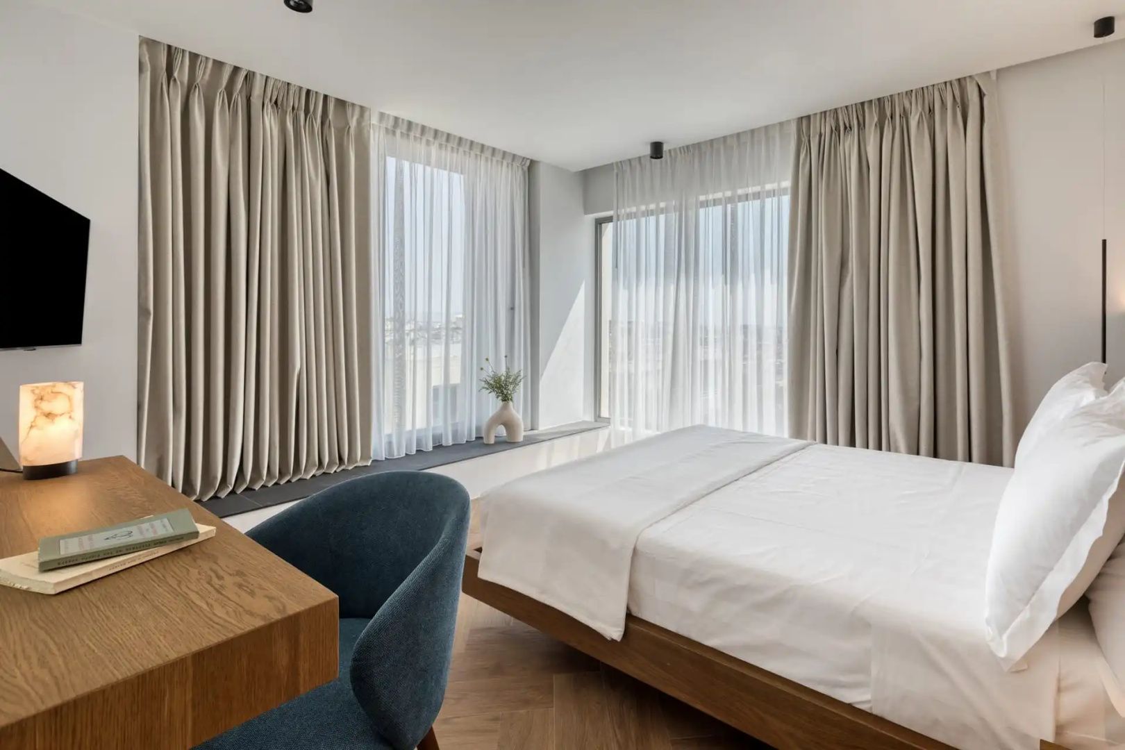 Schlafzimmer mit Doppelbett, Parkettboden, blauem Stuhl und bodentiefen Fenstern mit Gardinen.