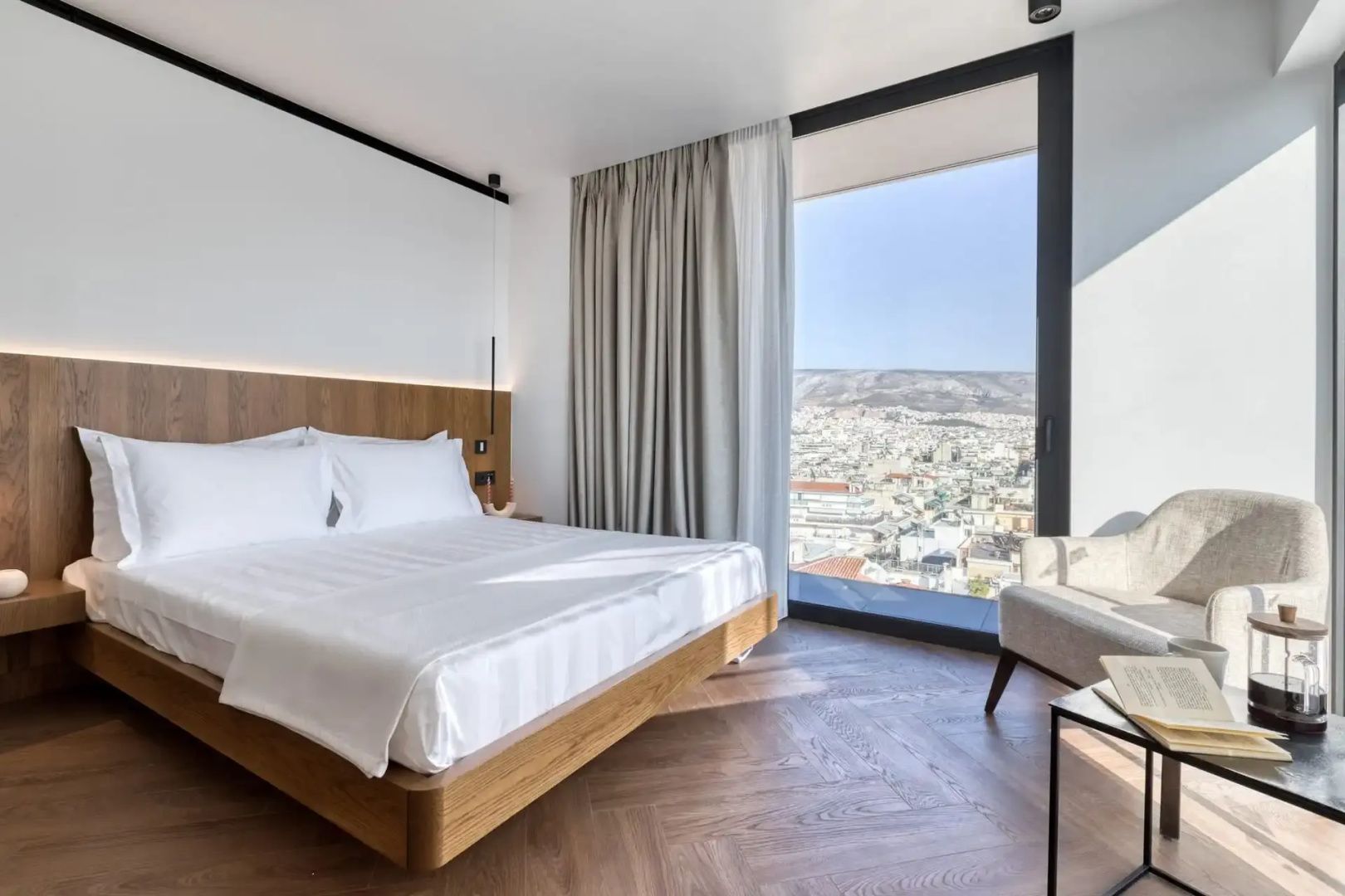 Doppelbett aus Holz in einem modernen Zimmer mit bodentiefem Fenster und Weitblick über die Stadt.