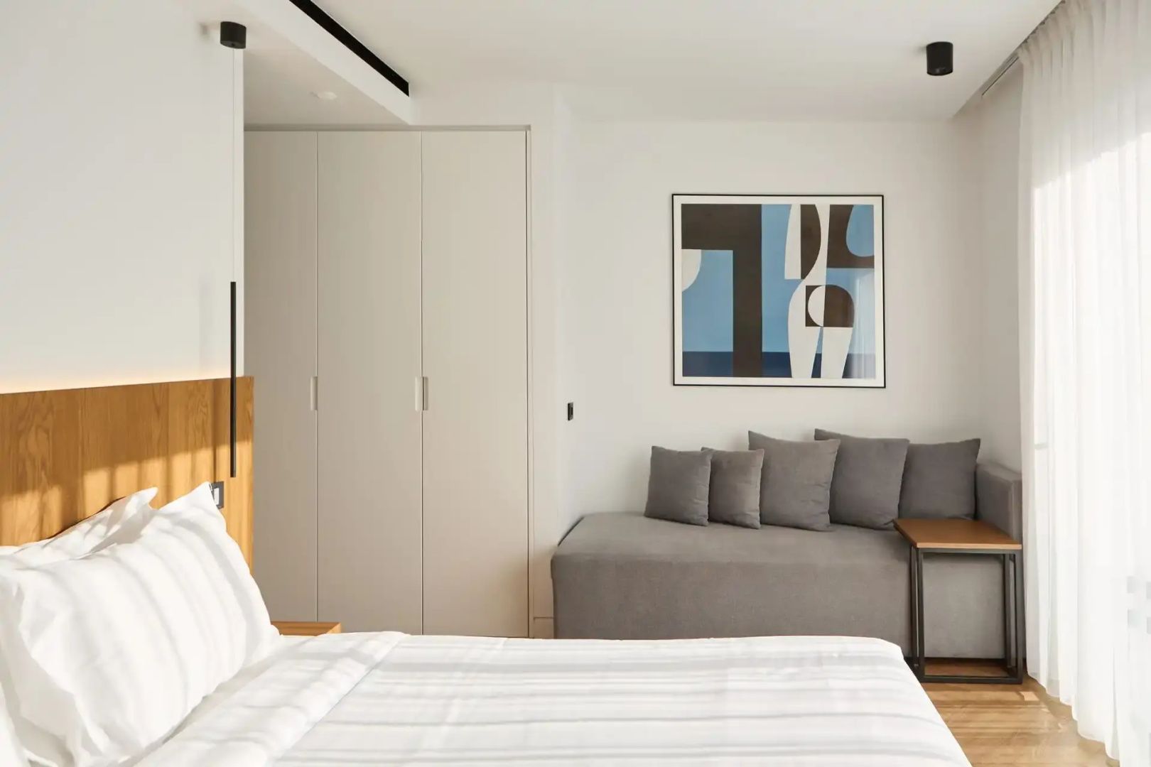 Hotelzimmer mit weißem Bett, grauer Schlafcouch, Einbauschrank und Kunstwerk.