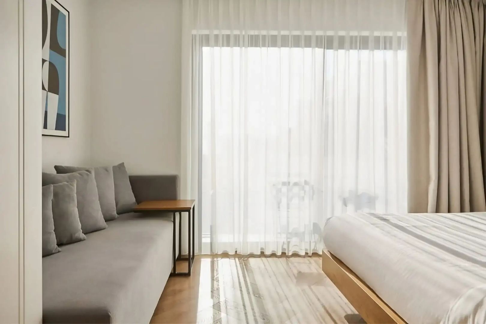 Helles Hotelzimmer mit grauem Sofa, Beistelltisch, Holzfußboden und Vorhang vor dem Fenster.