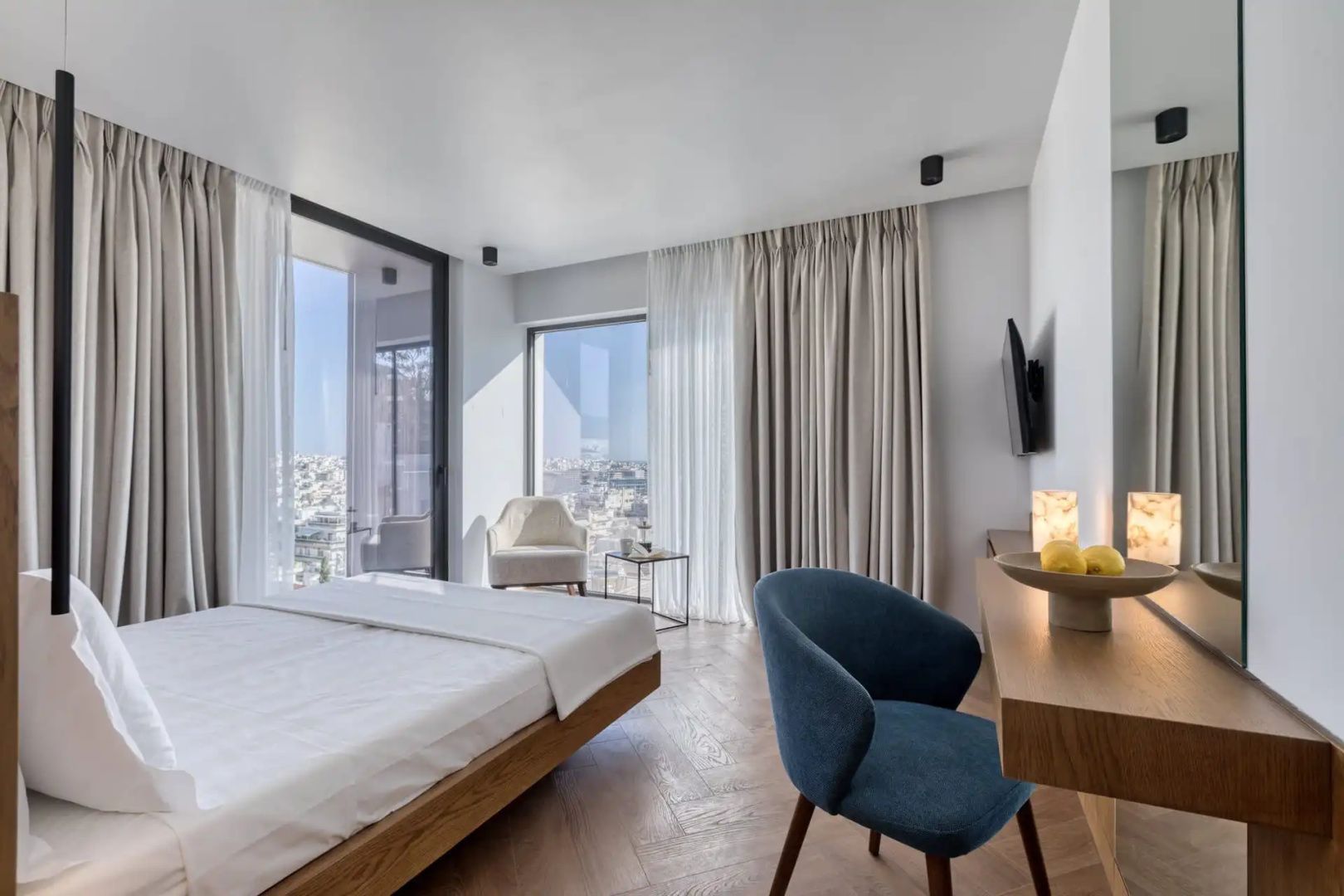 Helles Hotelzimmer mit Doppelbett, blauem Sessel, Holzboden und Ausblick auf die Stadt.