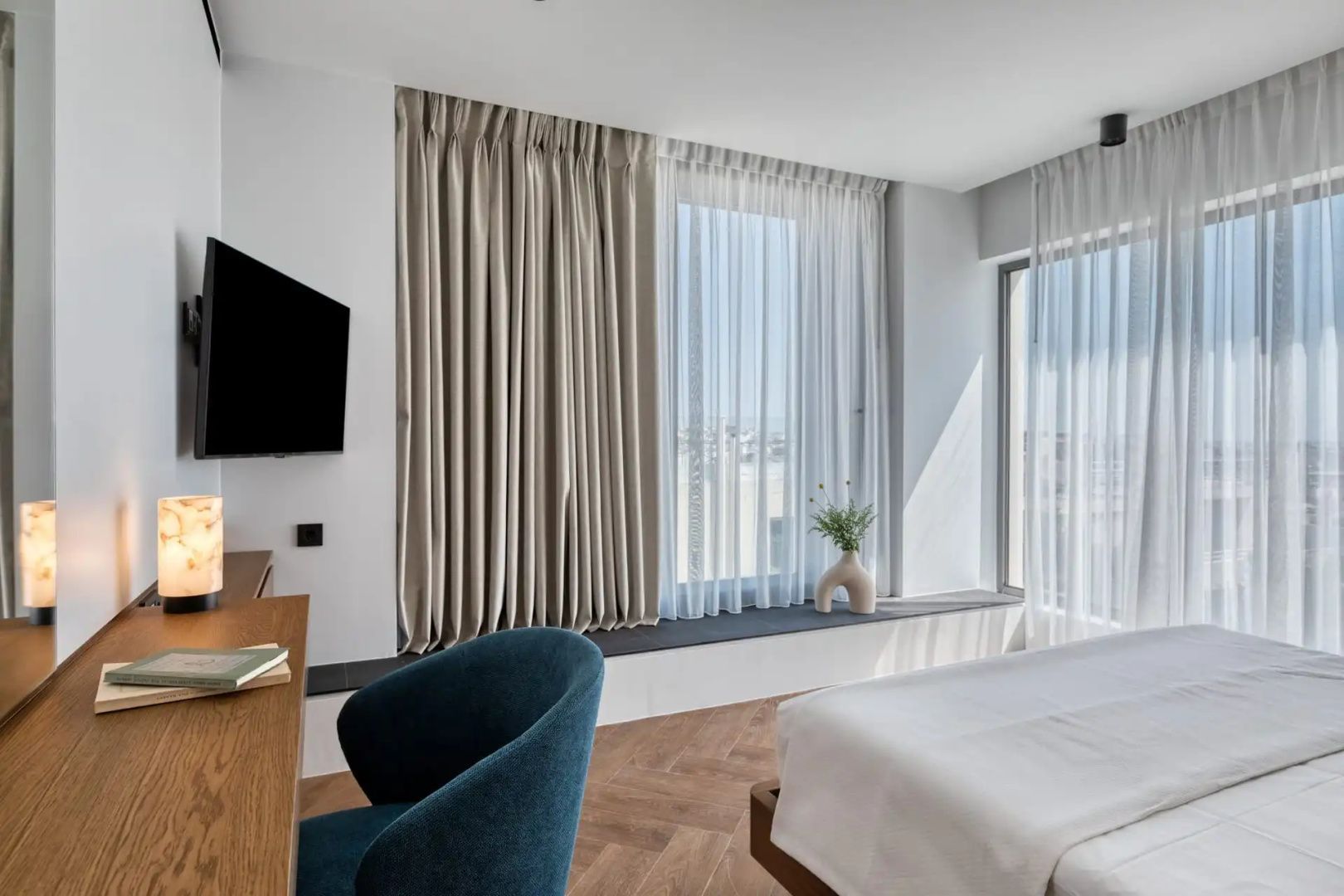 Helles Hotelzimmer mit Doppelbett, blauem Sessel, Schreibtisch und großem Fenster mit Vorhängen.