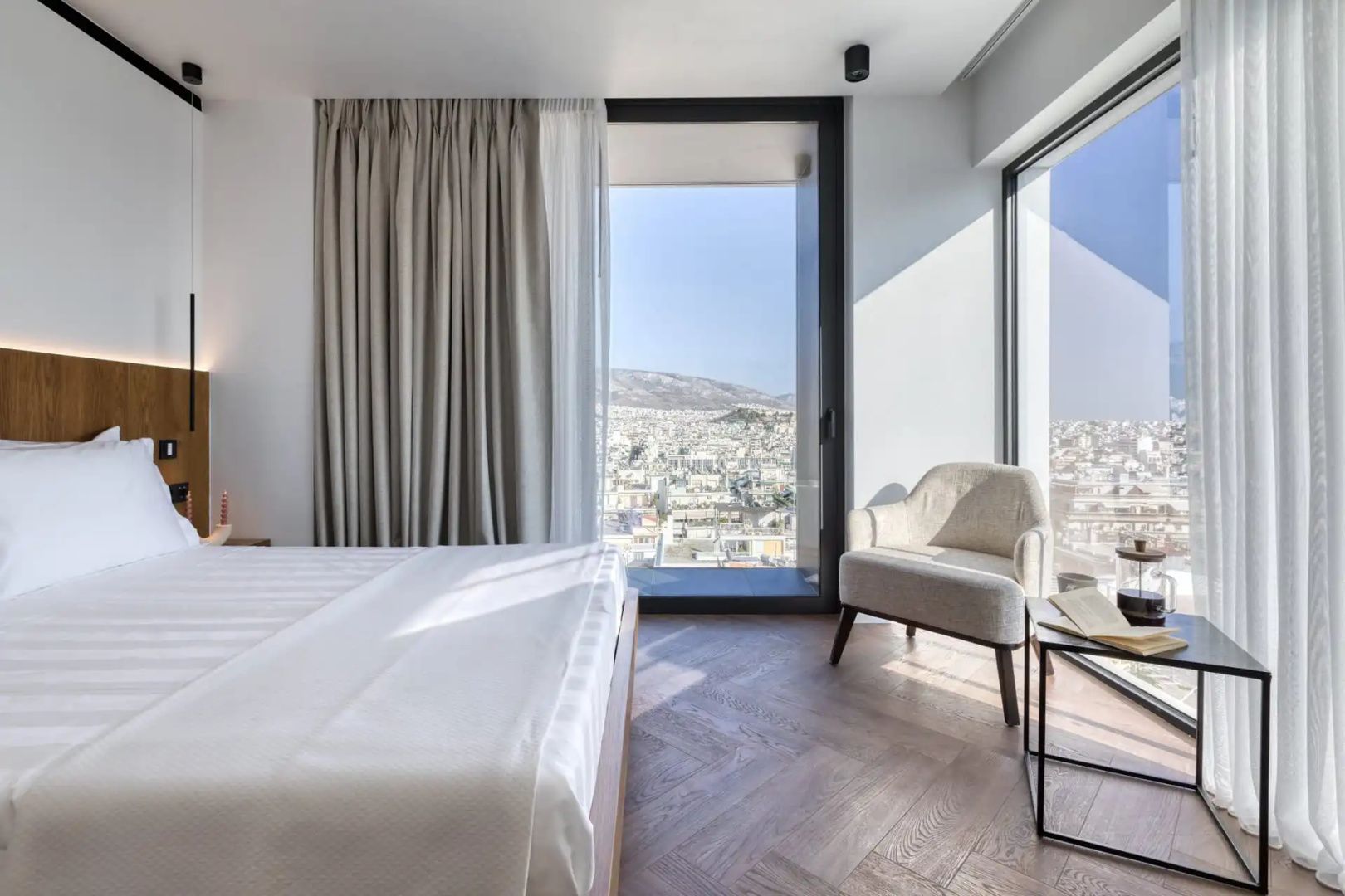 Modernes Hotelzimmer mit Parkettboden, weißem Bett, Sessel und Ausblick auf die Stadt.