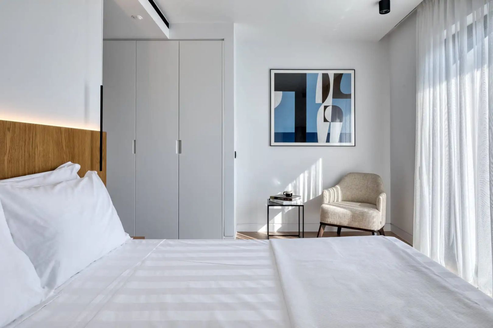 Modernes Hotelzimmer mit weiß bezogenem Bett, weißem Einbauschrank, beigem Sessel und Bild an der Wand.
