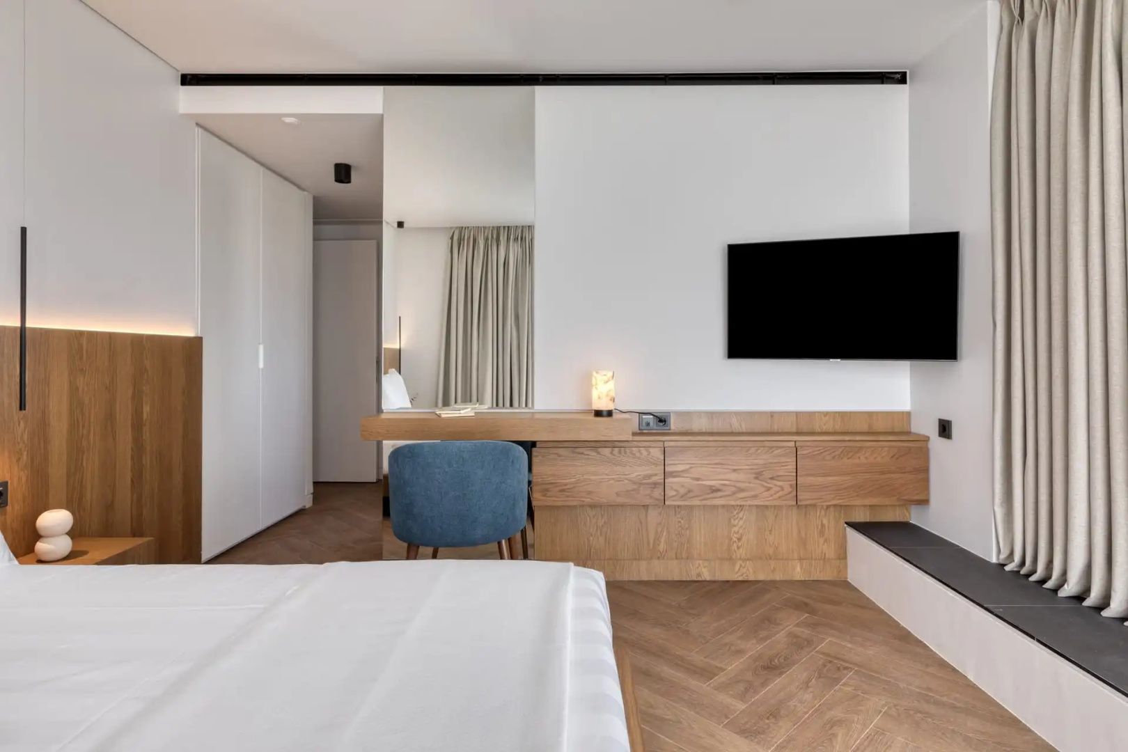 Hotelzimmer mit Holzmöbeln, wandmontiertem Fernseher, blauem Stuhl und weißem Kleiderschrank.
