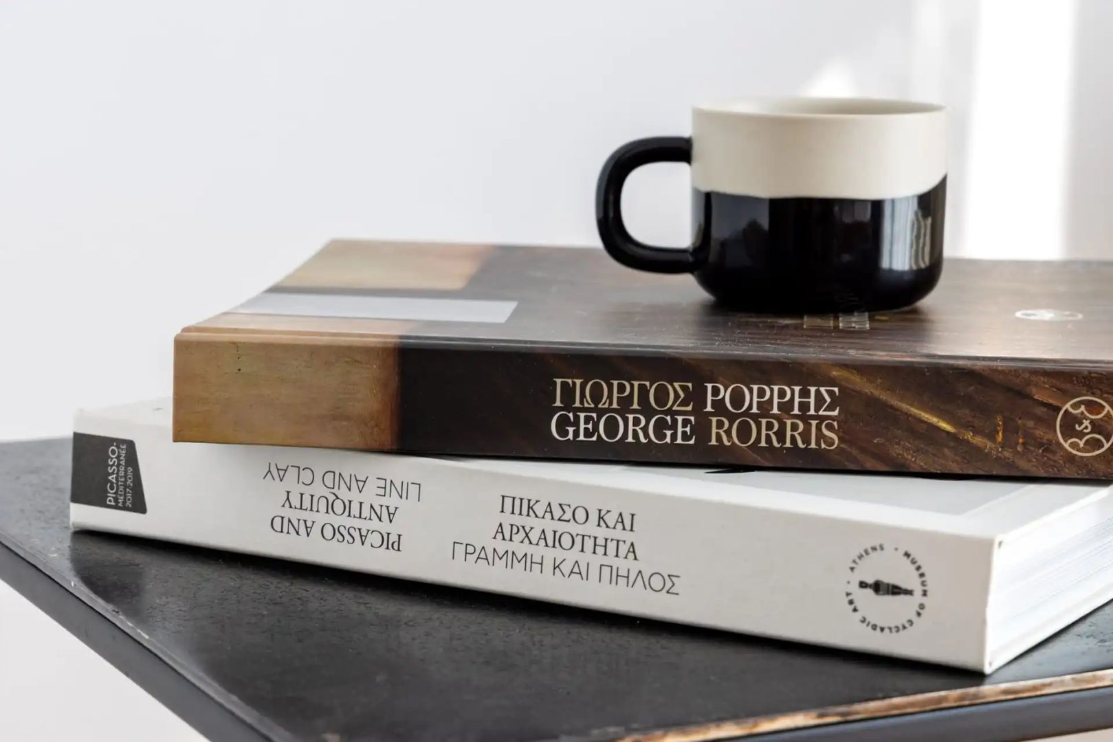 Zwei gestapelte Kunstbücher und eine Kaffeetasse auf einem dunklen Tisch.