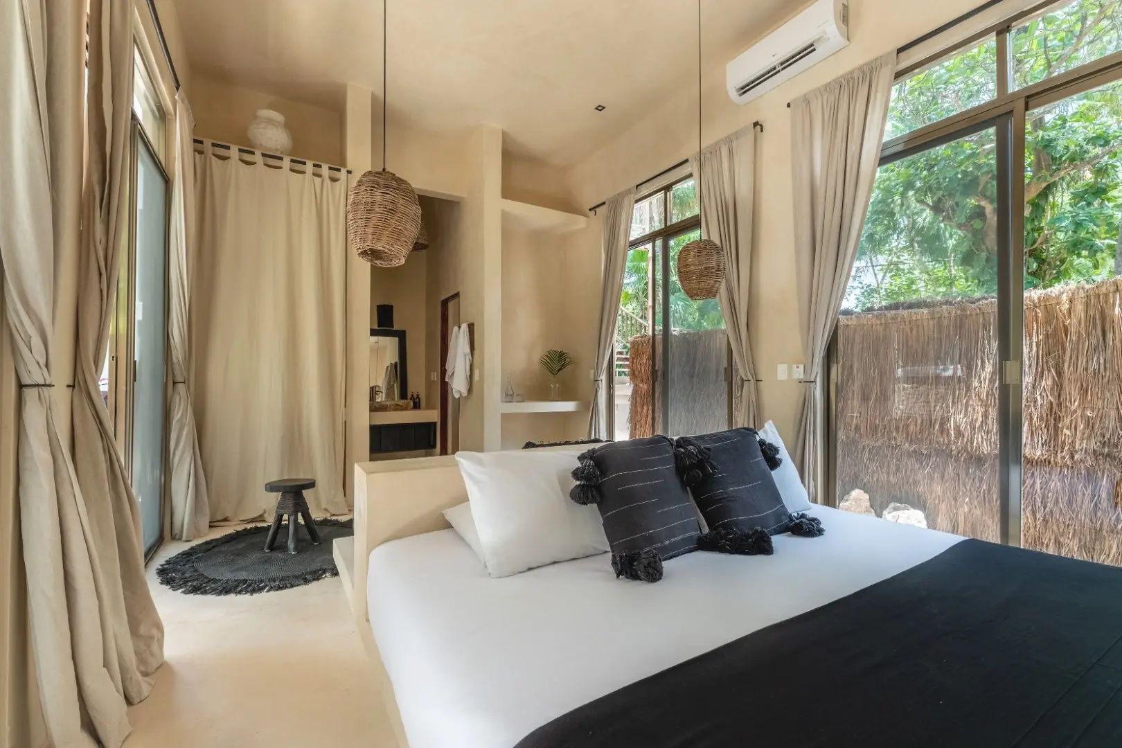 Bett mit weißen Kissen und dunkler Decke in einem hellen Zimmer mit Flechtlampen.
