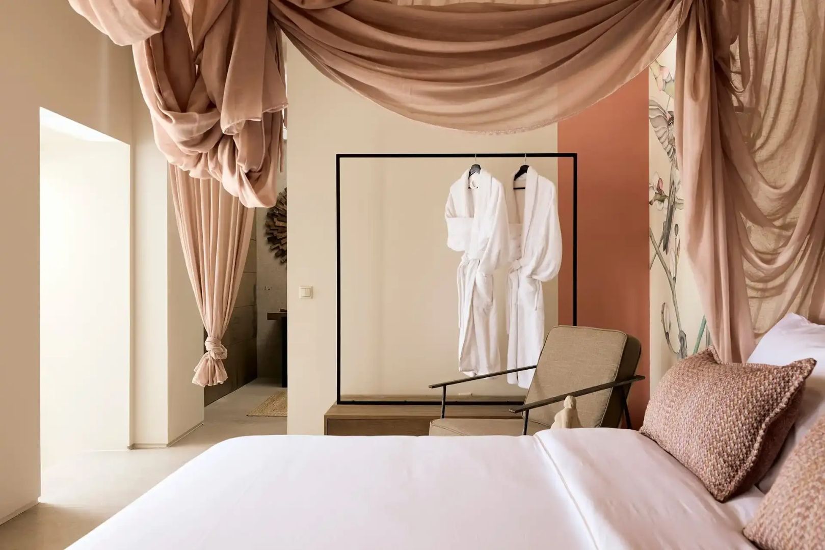 Helles Hotelzimmer mit Doppelbett, Stoffvorhängen und Kleiderständer mit Bademänteln.