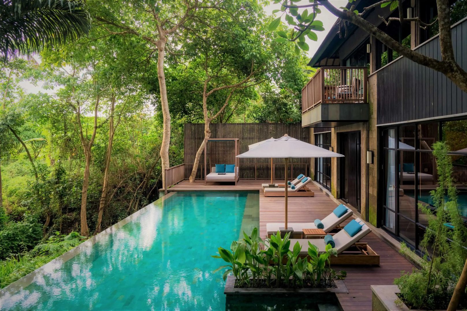 Poolbereich mit Sonnenliegen, Sonnenschirmen und Holzterrasse an einer Villa im Grünen.