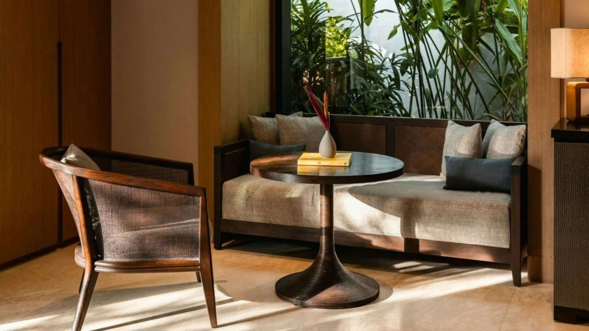 Sitzbereich im Zimmer mit rundem Holztisch, grauem Sofa und großen Fenstern ins Grüne.