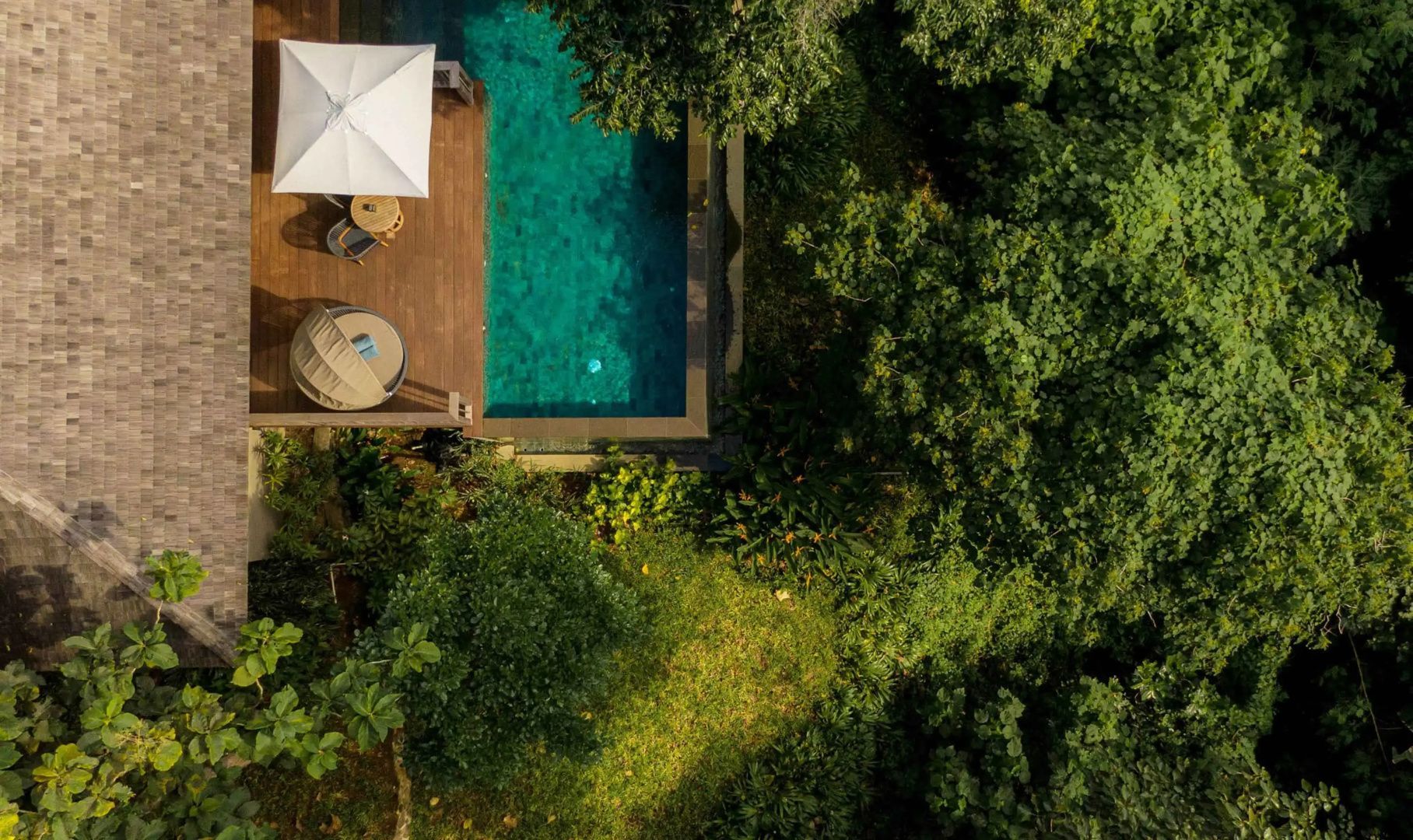 Draufsicht auf einen türkisblauen Swimmingpool mit Holzdeck inmitten dichter tropischer Bäume.