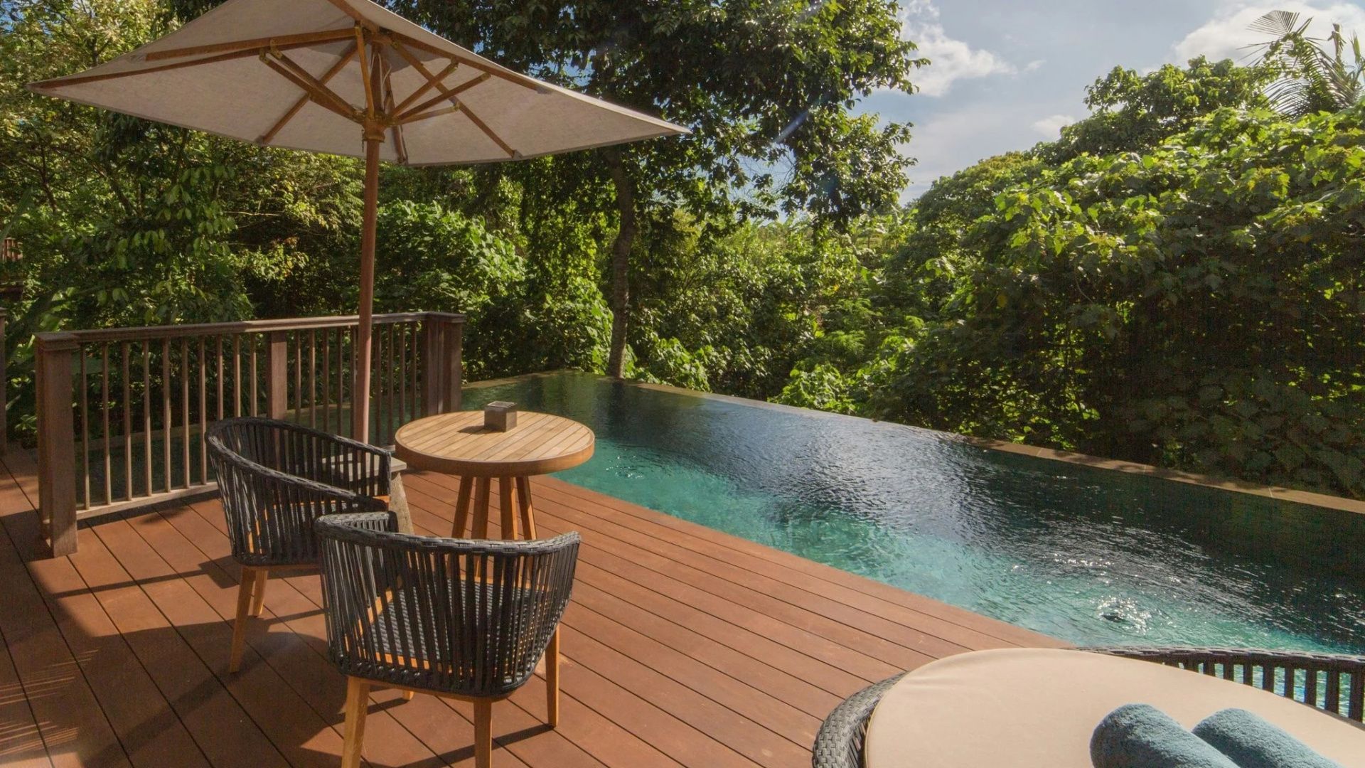 Holzdeck mit Sitzgruppe und Sonnenschirm an einem Infinity-Pool vor dichtem Wald.