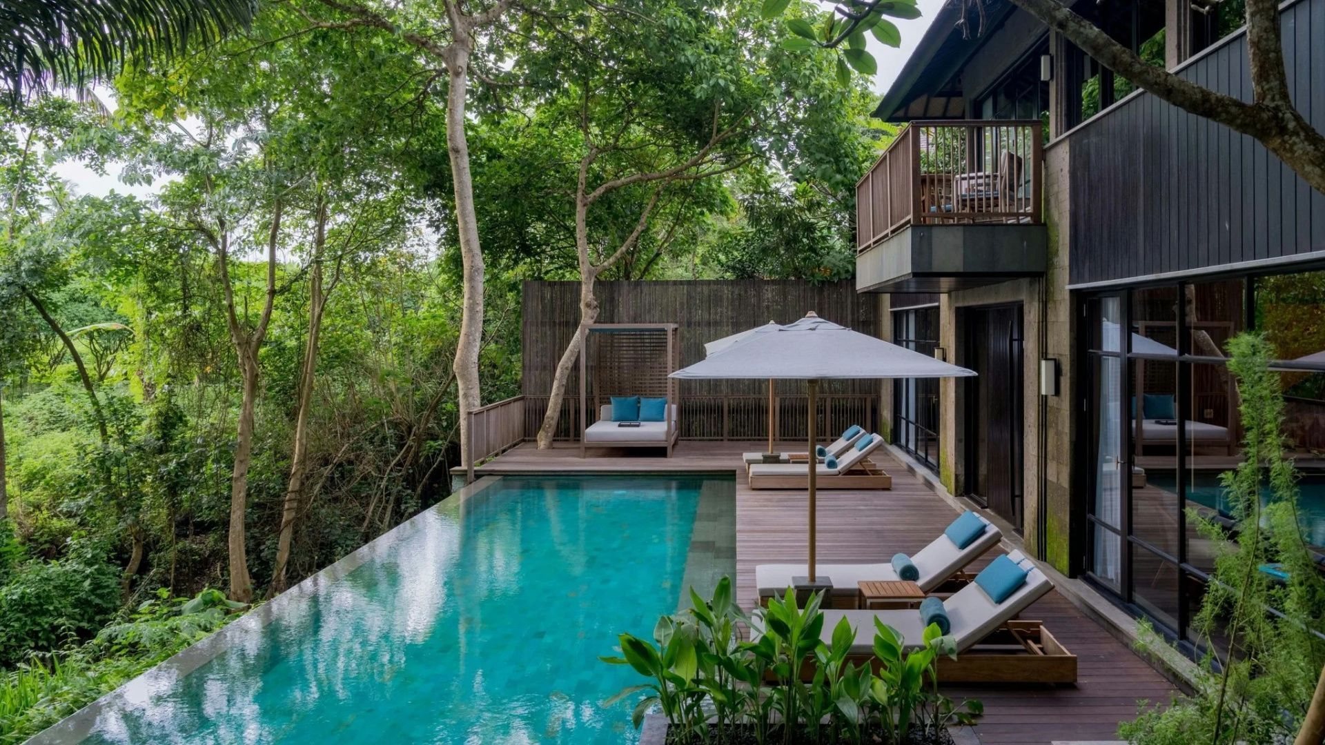 Privater Pool mit Holzdeck, Sonnenliegen und Sonnenschirm umgeben von dichten grünen Bäumen.