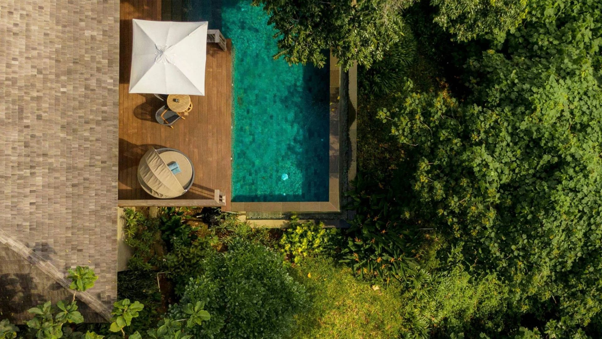 Vogelperspektive auf einen Pool mit Holzterrasse, Sonnenschirm und dichten tropischen Pflanzen.