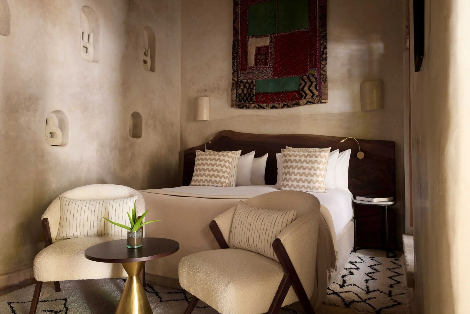 Doppelbett mit Holzkopfteil, beigen Kissen und zwei Sesseln an einem kleinen Tisch.