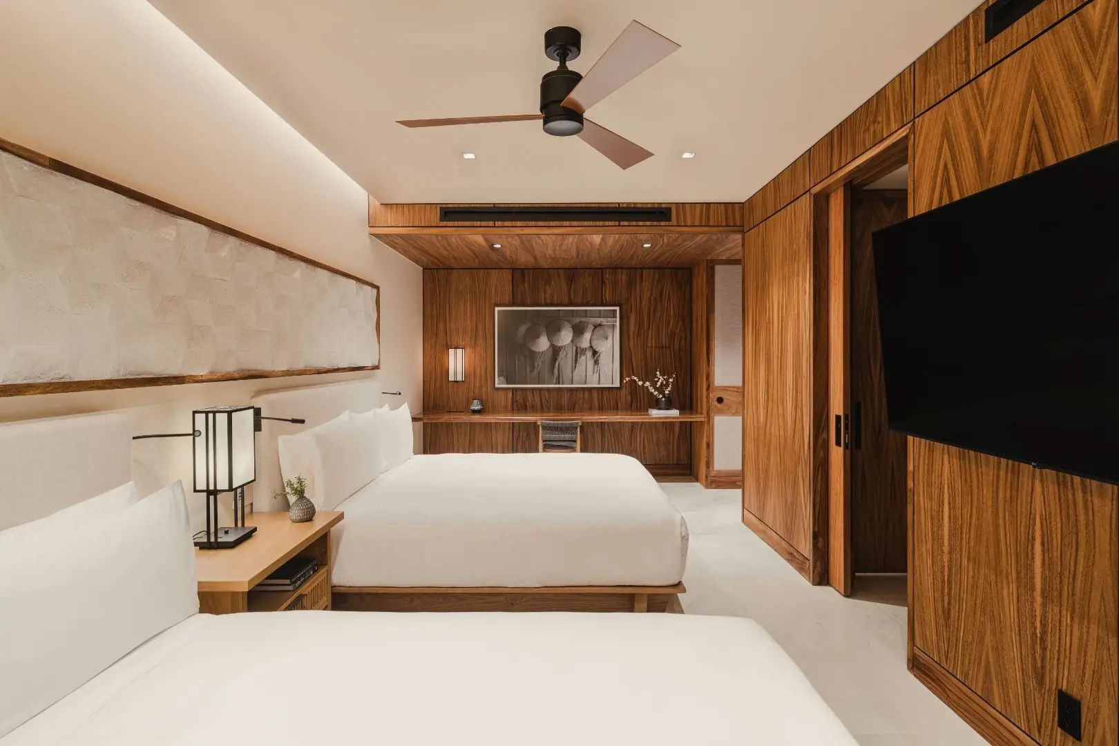 Helles Schlafzimmer mit zwei Betten, Deckenventilator und Schreibtisch vor einer Holzwand.