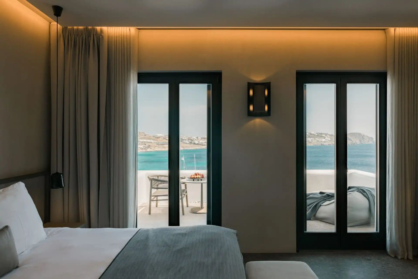 Blick aus dem Zimmer durch Glastüren auf eine Terrasse und das blaue Meer.