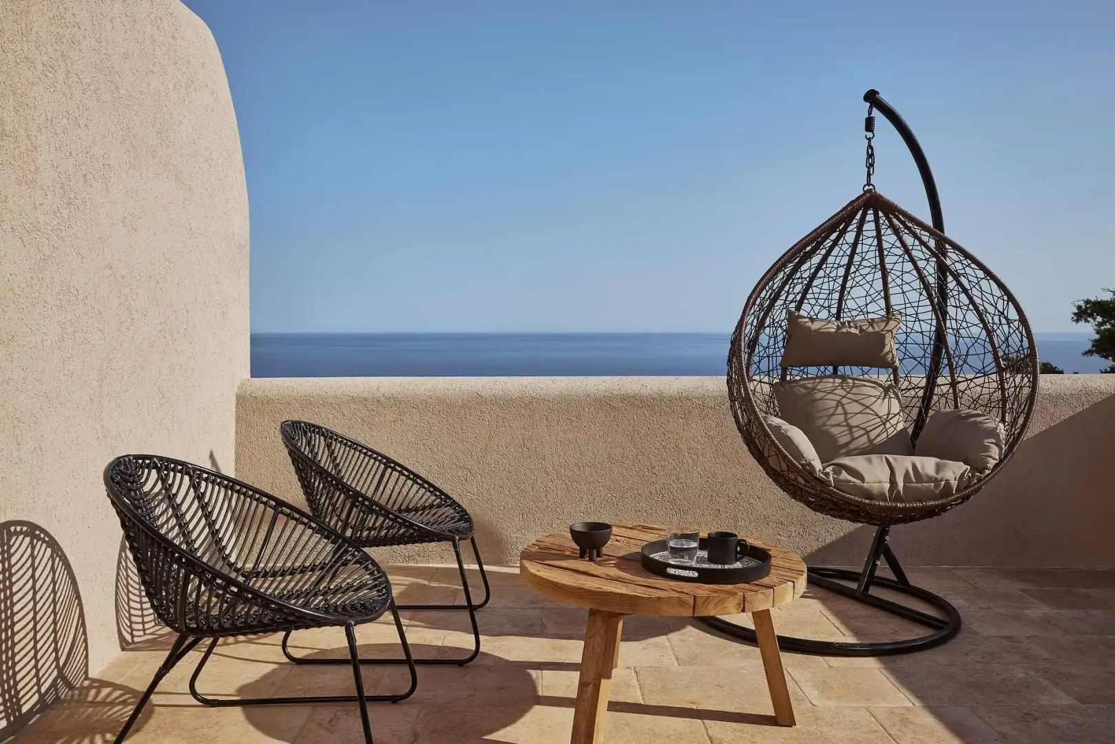 Terrasse mit zwei schwarzen Stühlen, Holztisch und Hängesessel vor dem blauen Meer.
