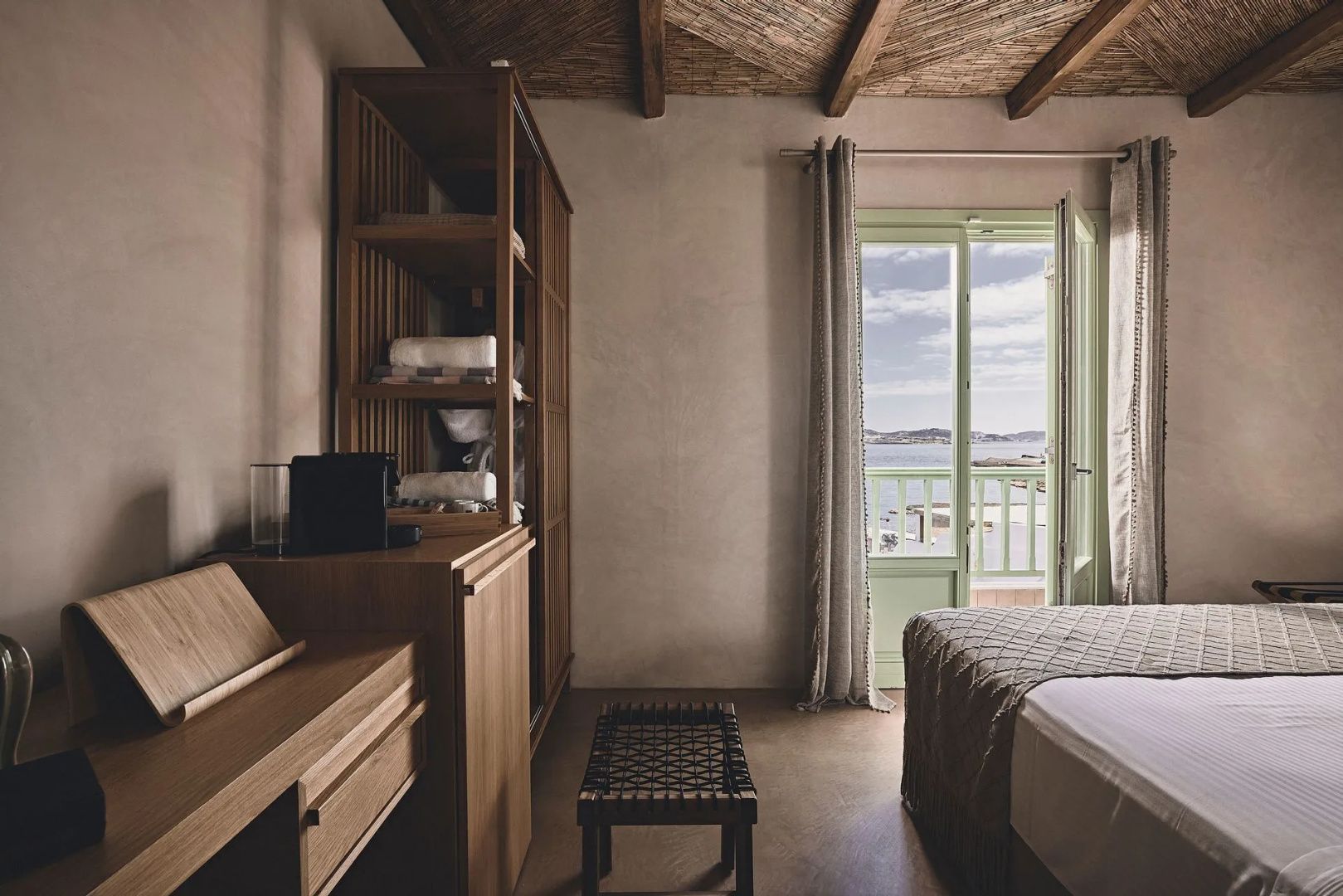 Schlafzimmer mit Holzschrank, Schreibtisch und geöffneter Balkontür mit Meerblick.