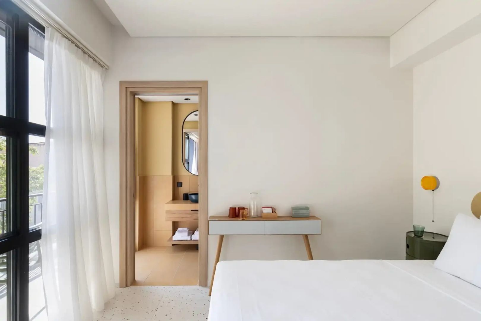 Heller Schlafraum mit Bett, Holzschreibtisch und Blick ins angrenzende Badezimmer mit gelber Wand.