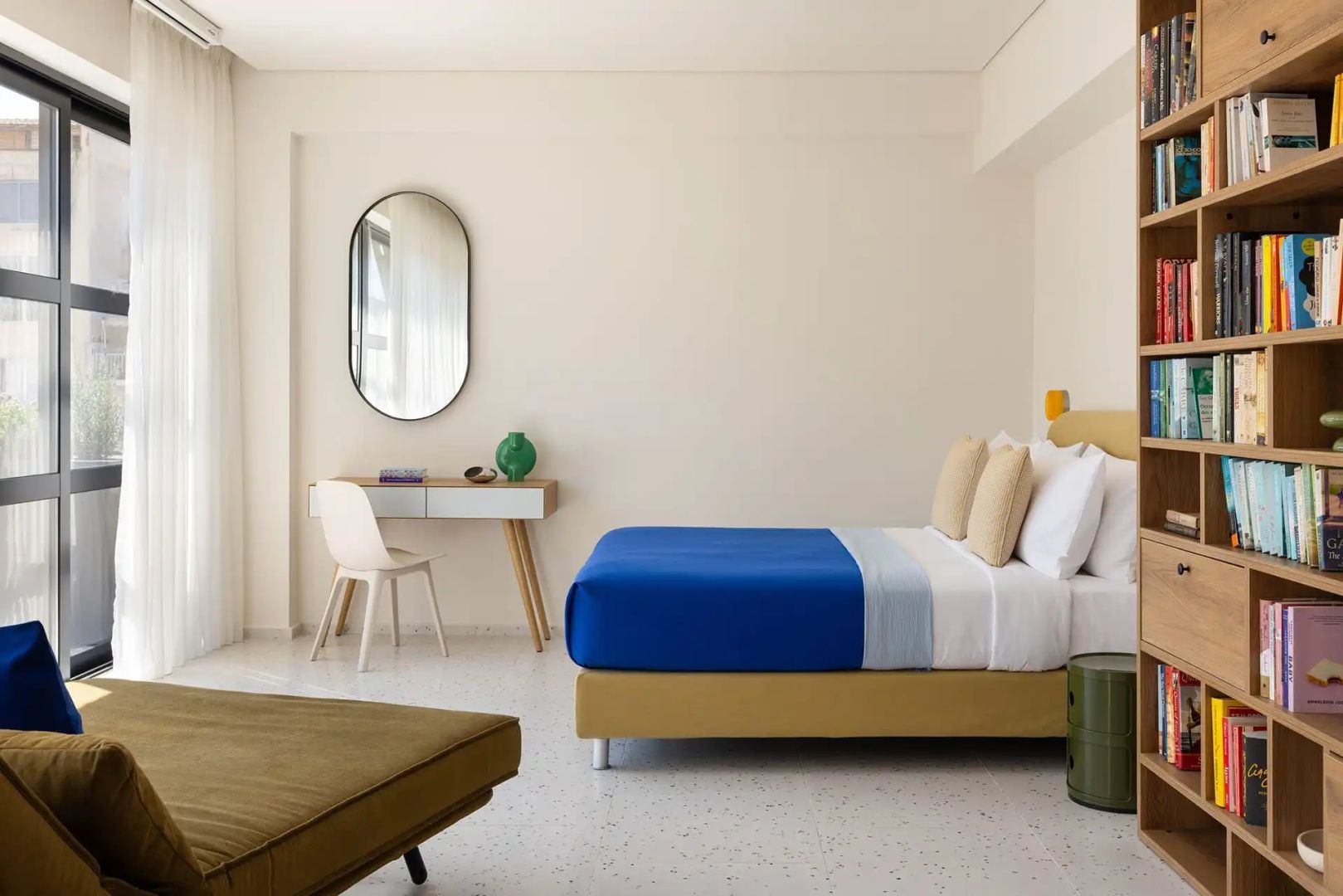 Helles Hotelzimmer mit blauem Bett, Schreibtisch, Stuhl und ovalem Wandspiegel.