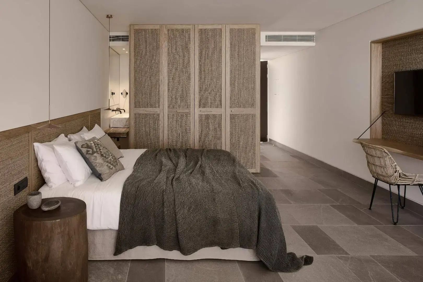 Doppelbett mit brauner Tagesdecke und dahinterstehendem Kleiderschrank mit Flechtwerk in einer Suite.